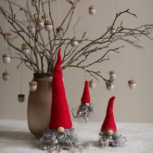 May include: Three felt gnomes with red hats and gray beards hanging from a branch with brown acorns. The gnomes are different sizes and are hanging from the branch with string.