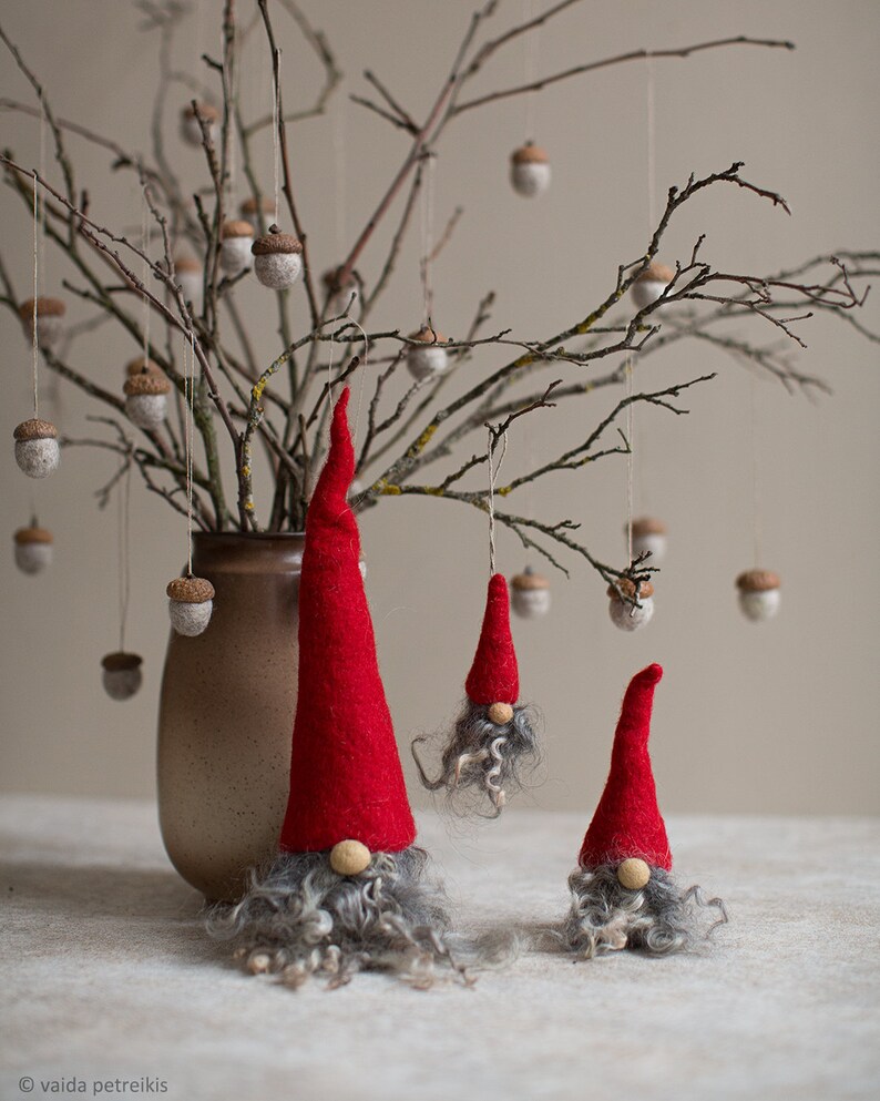 May include: Three felt gnomes with red hats and gray beards hanging from a branch with brown acorns. The gnomes are different sizes and are hanging from the branch with string.