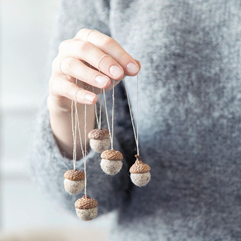 May include: Five brown felt acorns hanging from strings held by a hand. The acorns are a light brown color with a darker brown cap.