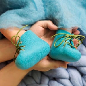 Puede incluir: Un par de botines de beb&eacute; de fieltro azul con cordones verdes. Los botines est&aacute;n atados con un lazo y se sostienen en la mano de una persona.