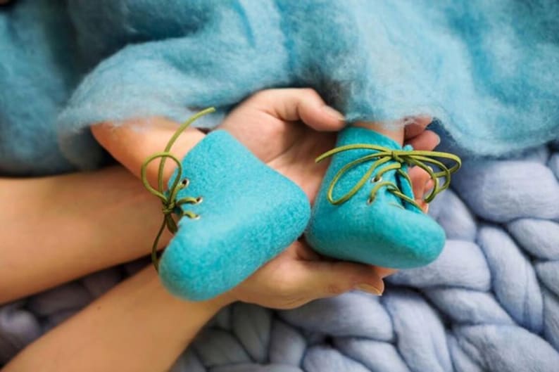 Puede incluir: Un par de botines de beb&eacute; de fieltro azul con cordones verdes. Los botines est&aacute;n atados con un lazo y se sostienen en la mano de una persona.