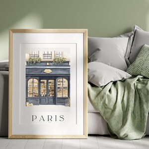 Op de afbeelding: Ingelijste print met een aquarel illustratie van een Parijse winkel met een blauwe gevel en gewelfde ramen. De print bevat het woord "PARIS" in een eenvoudig lettertype aan de onderkant. Het kunstwerk wordt gepresenteerd in een licht houten frame.
