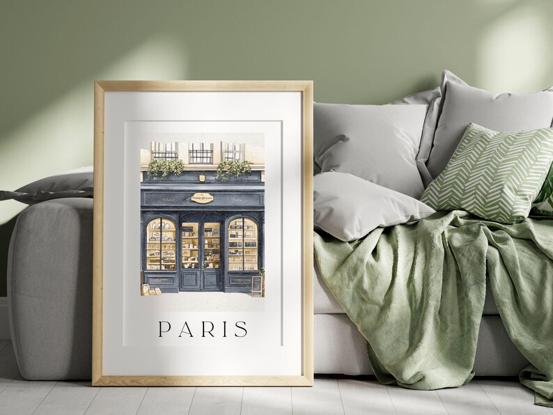 Op de afbeelding: Ingelijste print met een aquarel illustratie van een Parijse winkel met een blauwe gevel en gewelfde ramen. De print bevat het woord "PARIS" in een eenvoudig lettertype aan de onderkant. Het kunstwerk wordt gepresenteerd in een licht houten frame.