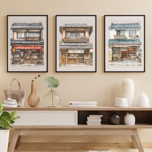 Puede incluir: Tres acuarelas enmarcadas de fachadas de tiendas japonesas con marcos negros. Las pinturas est&aacute;n colgadas en una pared beige sobre un estante de madera con art&iacute;culos decorativos.