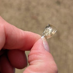 May include: A small, clear, rectangular pendant with a silver-colored wire. The pendant has a light amber hue and contains a small, abstract design. The pendant is held between two fingers against a blurred, neutral background.