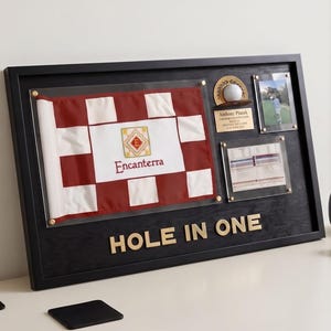 May include: A black wooden display frame featuring a red and white checkered golf flag with the text Encanterra, a golf ball, a personalized plaque, a photograph, and a scorecard. The words HOLE IN ONE are mounted in gold lettering at the bottom.