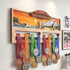 May include: A wooden medal display rack featuring a sunset landscape with cutouts of a swimmer, cyclist, and runner. The top banner reads ANTHONY and FAILURE IS WHEN YOU STOP TRYING. Multiple colorful ribbons with various metal medals hang below.