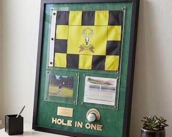 Puede incluir: Un marco negro con fondo verde que contiene una bandera de golf a cuadros amarillos y negros con un logotipo de cráneo de ciervo, una fotografía, una tarjeta de puntuación, una pelota de golf y una placa dorada con el texto HOLE IN ONE.