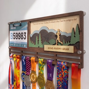 Op de afbeelding: Een houten medaillehouder met een clip voor een startnummer met het nummer B59983, een gelaagde uitgesneden berglandschap en de tekst "The miracle isn't that I finished. The miracle is that I had the courage to start" en "ANNE-MARMY-GRAM".
