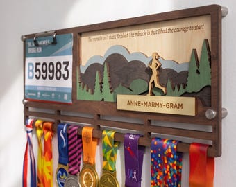 May include: A wooden wall-mounted medal display rack featuring a clip for a race bib with the number B59983, a layered mountain and forest landscape cutout, and the text "The miracle isn't that I finished. The miracle is that I had the courage to start" and "ANNE-MARMY-GRAM".
