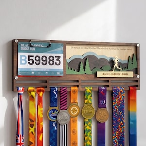 May include: A wooden medal display rack featuring a clip for a race bib with the text Blackmores Bridge Run B59983, a layered mountain landscape cutout, and the name Anne-Marmy-Gram. Nine colorful ribbons with various medals hang below.