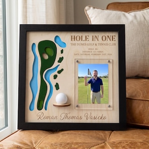 Puede incluir: Un marco de madera para pared con un mapa de campo de golf cortado a láser, una pelota de golf blanca en un estante pequeño y una foto de una persona en un campo de golf. El texto dice: HOLE IN ONE, THE DUNES GOLF & TENNIS CLUB, HOLE: #6.