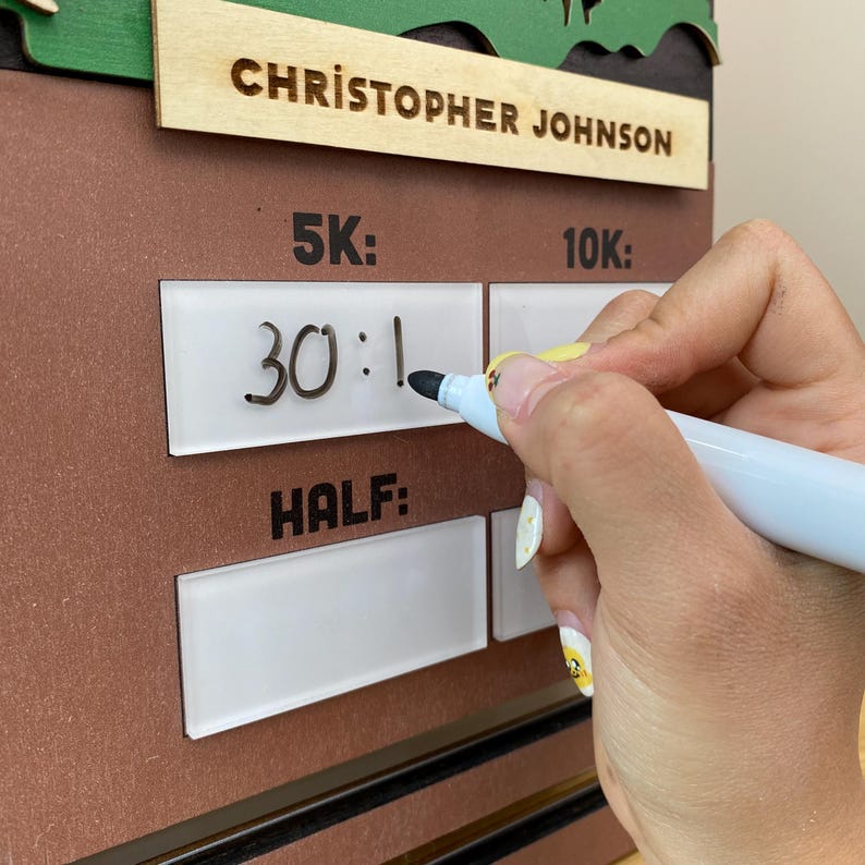 May include: A brown wooden race tracker board with the name CHRISTOPHER JOHNSON engraved at the top. The board features white rectangular sections labeled 5K, 10K, and HALF for recording race times with a white dry-erase marker.