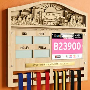 May include: Wooden medal display with a cityscape design, the word "FINISH", and the name "JAMES THOMPSON". Includes slots for race distances and a pink race bib with the number B23900. Features medal ribbons.
