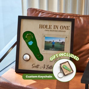 May include: A wooden golf-themed display with the text "HOLE IN ONE" and "OMNI BEDFORD SPRINGS". It includes a photo, a golf ball, and a green golf course cutout. The text "GIFT INCLUDED" and "Custom Keychain" are also visible.