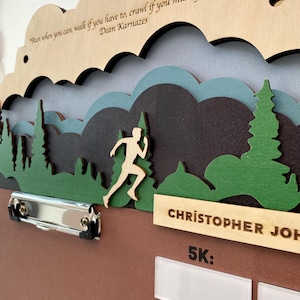 May include: A wooden medal display board featuring a layered mountain and forest landscape with a runner silhouette. The text reads: Run when you can, walk if you have to, crawl if you must, Dean Karnazes, CHRISTOPHER JOH, and 5K.