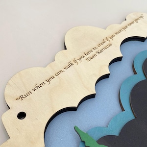 May include: A layered wooden wall hanging featuring a light wood top layer with the engraved text "Run when you can, walk if you have to, crawl if you must, just never give up" by Dean Karnazes, with blue, green, and black cloud-shaped layers.
