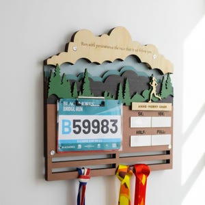 May include: A wooden race medal and bib display rack featuring a mountain and forest silhouette, a clip for a race bib with the text B59983, and sections for race times labeled 5K, 10K, HALF, and FULL. The top includes the text Run with perseverance the race that is set before you.