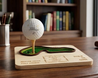 Può includere: Un accessorio da scrivania a tema golf con una pallina da golf su un tee, posizionata sopra un mini green. La base in legno è incisa con "Hole in One" e "Beaver Creek Golf Course".
