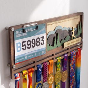 May include: A wooden wall-mounted medal display rack featuring a metal clip holding a Blackmores Bridge Run race bib number B59983. The display includes a layered wooden mountain scene, the text "The miracle isn't that I finished. The miracle is that I had the courage to start," and a name plate reading "ANNE-MARMY-GROM" with multiple colorful ribbons hanging below.