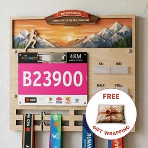 Puede incluir: Un expositor de madera para dorsales y medallas con un paisaje de montaña, un clip metálico que sujeta un dorsal rosa con el texto B23900 y ranuras para colgar medallas. La pancarta superior muestra el texto NATHAN'S MEDAL.