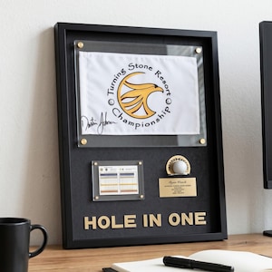 Könnte beinhalten: Eine gerahmte Golf-Erinnerungsanzeige mit einer weißen Flagge mit dem Logo der Turning Stone Resort Championship, einer Scorekarte, einem Golfball und einer Plakette. Die Worte "HOLE IN ONE" sind in goldenen Buchstaben dargestellt.