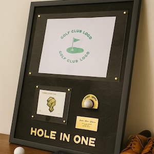 May include: A framed golf achievement display with a black frame. The display includes a golf club logo, a small framed image, a golf ball with a gold accent, and a plaque that reads "HOLE IN ONE". A golf ball, glove, and brown leather shoes are next to the frame.