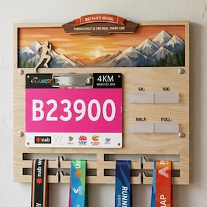 May include: Wooden medal display board with a mountain scene, a runner silhouette, and the text "Nathan's Medal." Includes a pink race bib, medal ribbons, and spaces for race distances. The text "Consistency is the real finish line" is also visible.