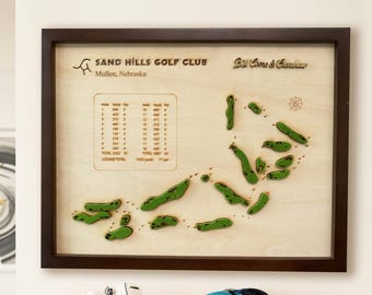 May include: A framed wooden golf course map of Sand Hills Golf Club in Mullen, Nebraska. The map features green course layouts on a light wood background, with the text "Sand Hills Golf Club" and "Bill Coore & Crenshaw". The frame is dark brown.