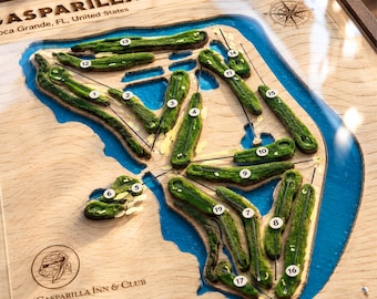May include: A wooden 3D topographic map of the Gasparilla Golf Club in Boca Grande, FL, featuring green textured fairways, blue water hazards, and numbered holes from 1 to 19. The map includes a compass rose and The Gasparilla Inn & Club logo.