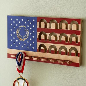 Könnte beinhalten: Medaillen-Display aus Holz im Stil der amerikanischen Flagge. Es hat einen blauen Bereich mit weißen Sternen und einem goldenen Kranz, rote und weiße Streifen und gewölbte Schlitze für Medaillen. Eine Baseball-Medaille hängt unten.