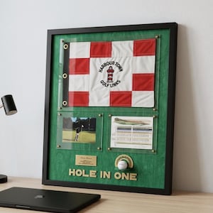 May include: Framed golf display with a red and white chequered flag, the text "Harbour Town Golf Links", photos, a scorecard, and a golf ball. The text "Hole in One" is displayed. The frame is black with a green background.