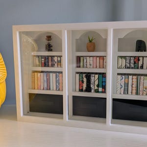 May include: A white miniature bookshelf with three sections, each containing small, colorful books. The shelves also hold decorative items like a small plant and a dark figurine. A large yellow owl figurine is on the left.