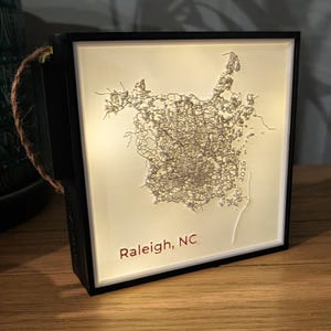 May include: A square, black-framed, illuminated light box featuring a 3D-printed, cream-colored relief map of Raleigh, NC. The map includes intricate street networks, the text 'Raleigh, NC' in the bottom left, and the year '2026' on the right side.