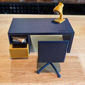 May include: A miniature dark blue desk with a yellow drawer and a matching yellow desk lamp on top. The desk features a small open shelf containing a book and the text SappySlabs printed on the surface. A dark blue chair with a tripod base sits in front.