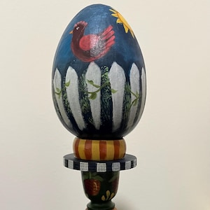May include: A decorative wooden egg-shaped stand with a painted farm scene. The egg features a red chicken, a white picket fence, and a yellow sun against a blue background. The stand has a black and white checkered base and a sunflower design.