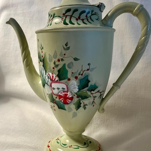 May include: A light green, hand-painted coffee pot with a red finial and a decorative handle. The pot is adorned with holly, red berries, and candy cane accents, perfect for holiday decor. The base has a red and green trim.