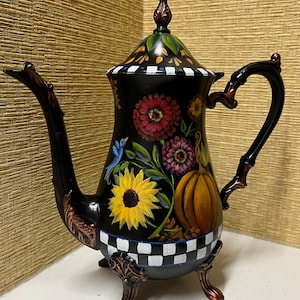 May include: A black decorative teapot with a floral and pumpkin design. The teapot features a sunflower, a blue bird, and a checkered pattern. The handle and spout have bronze-colored accents.
