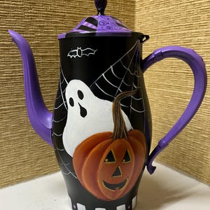 May include: A black Halloween teapot with a purple handle and spout. The teapot features a white ghost, an orange jack-o'-lantern, and a spider web design. The lid is decorated with purple and black stripes.