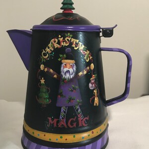 May include: Black metal Christmas-themed teapot with a purple lid and handle. The teapot features a painted Santa Claus figure holding a Christmas tree and a monkey. The word "CHRISTMAS" arches above Santa, and "MAGIC" is written below.