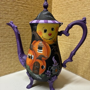 May include: A decorative teapot featuring a black base with hand-painted orange pumpkin houses, a yellow moon face, and a small spider wearing a purple hat. The teapot has a purple handle, spout, and feet, with a black lid topped by a purple finial.