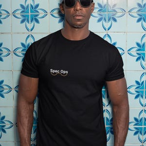 Könnte beinhalten: Ein Mann mit schwarzer Sonnenbrille und einem schwarzen T-Shirt mit dem Text "Spec Ops" und einer Brillengrafik. Er steht vor einer blauen und weißen gekachelten Wand.