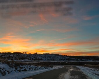May include: A scenic view of a snow-covered road winding through a mountainous landscape. The sky is a vibrant orange and pink, creating a beautiful sunset backdrop.