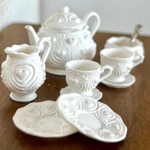 May include: White porcelain tea set featuring a heart design. The set includes a teapot, creamer, two cups, two saucers, and a plate.