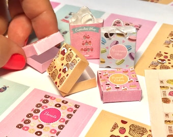 May include: Miniature bakery-themed packaging, including pink boxes and paper bags. The boxes feature patterns of pastries and cupcakes. The bags are printed with "Cupcakes Shop" and "Macarons Shop" text. The scene includes printed cards with donut and pastry designs.