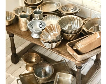 May include: A collection of miniature metal kitchenware in gold and silver tones. The set includes various baking pans, a colander, and a strainer. The items are arranged on a wooden table with a white background.