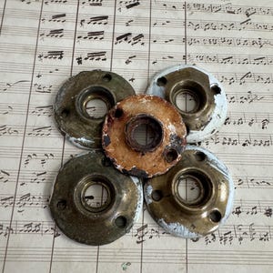 May include: Five circular metal hardware plates arranged in a cluster. Four plates feature a brass-toned finish with remnants of white paint around the edges, while the central plate is smaller, circular, and shows a rusted, brown-toned finish.