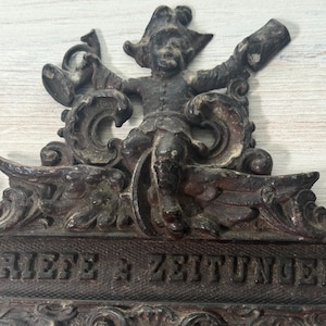May include: A dark brown, ornate metal wall plaque featuring a central figure in a hat holding a horn and a rectangular object. The plaque displays the embossed text "BRIEFE & ZEITUNGEN" across a textured horizontal bar with decorative scrollwork.