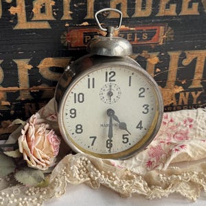 Puede incluir: Reloj despertador Majestic plateado vintage con esfera crema, números negros y campana. El reloj marca aproximadamente las 5:30. Una rosa seca y un ribete de encaje en primer plano, con una caja de madera envejecida al fondo.