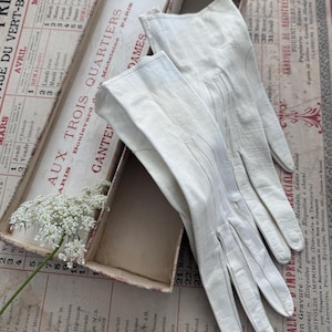 May include: A pair of off-white leather gloves with stitched detailing on the back of the hands, resting inside an elongated rectangular cardboard box labeled with the text Aux Trois Quartiers Ganterie Paris.